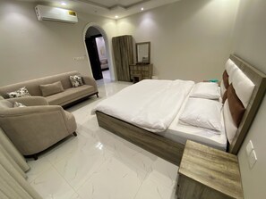 A modern hotel room with a large bed, a sofa, a wooden nightstand, and an air conditioning unit on the wall.