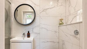 Un baño con pared de mármol, un espejo redondo, un lavabo con una botella de jabón y una zona de ducha.