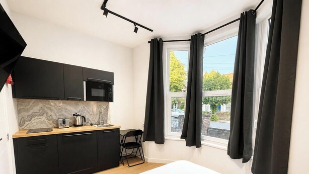A compact room with a kitchenette, a bed, a window with curtains, and a view of the outside. - Central Studio in the Heart of Hounslow! (Greater London)