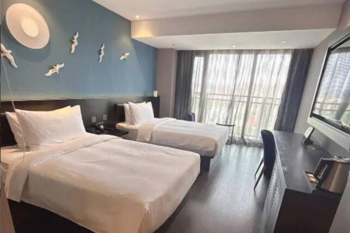 Habitación de hotel con dos camas, un escritorio, un televisor y un ventanal grande con cortinas.