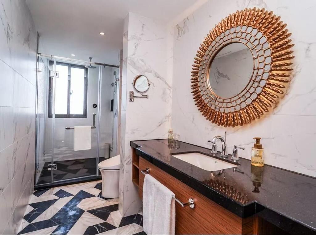 Un baño moderno con ducha de vidrio, suelo de mármol, lavabo con encimera negra y un espejo decorativo.