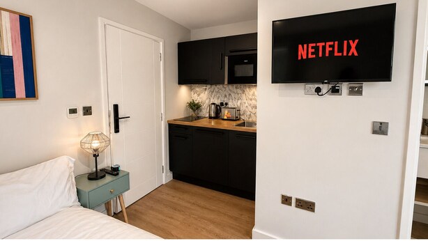 A modern hotel room with a bed, bedside table, lamp, kitchenette, and a TV displaying 'NETFLIX'. - Unique Studio in the Heart of Hounslow! (Greater London)