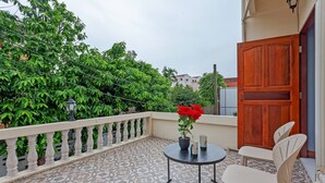 Een balkon met een betegelde vloer, een bloempot met rode bloemen, twee witte stoelen en een houten deur.