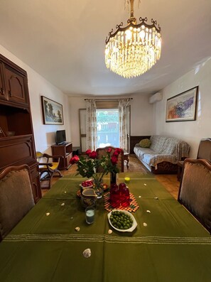 A dining area with a table set for two, a chandelier, and a view through a window.