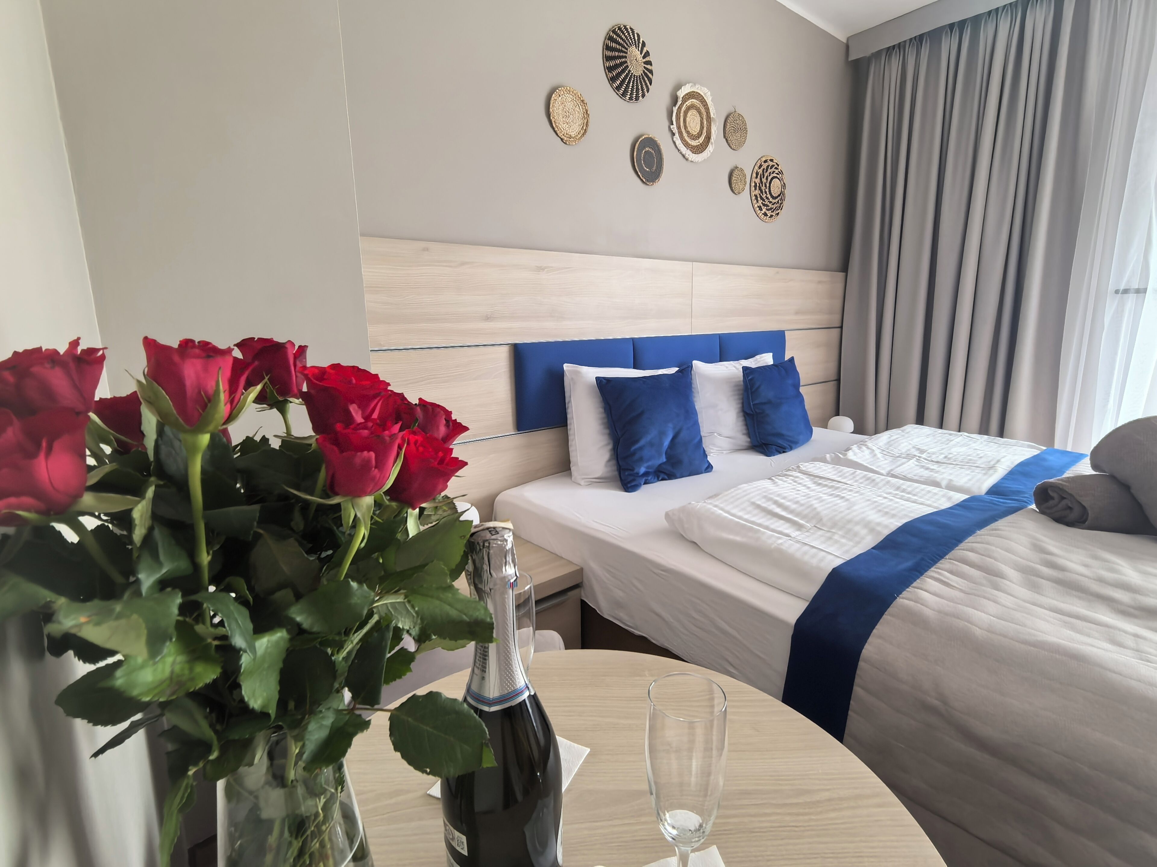 Ein Hotelzimmer mit Bett, Tisch mit Flasche und Glas sowie einer Vase mit roten Rosen.