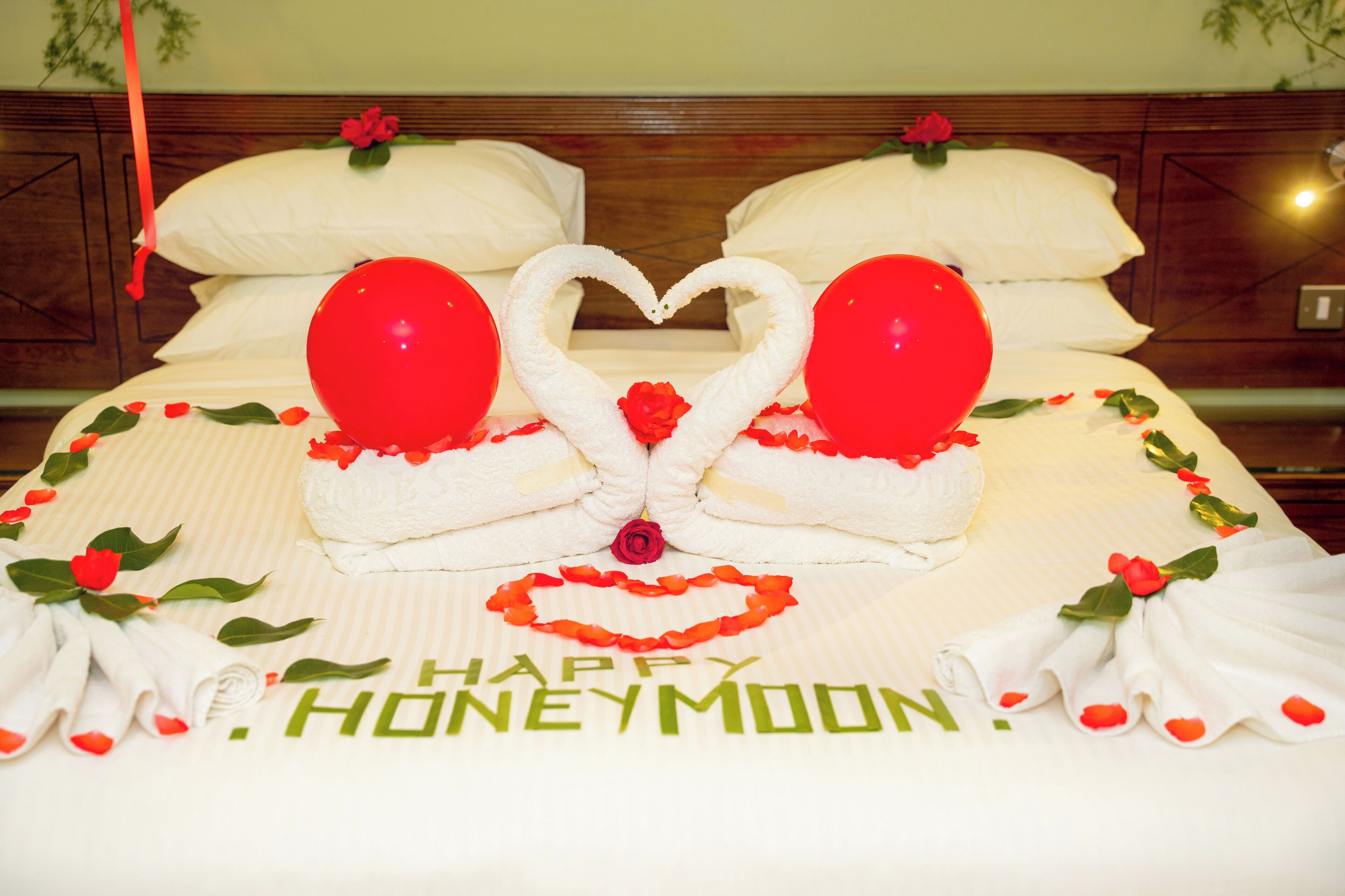 Cama com arranjo de balões em formato de coração, uma rosa e a palavra 'HONEYMOON' bordada no lençol.