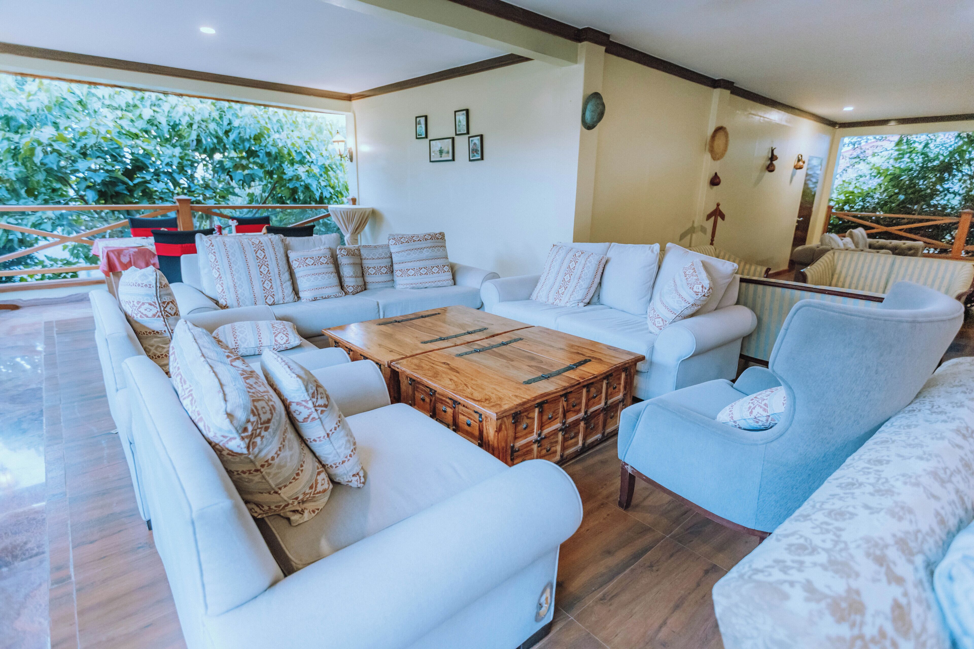 A spacious living area with a large wooden coffee table, white sofas with patterned cushions, and a view of greenery through large windows.
