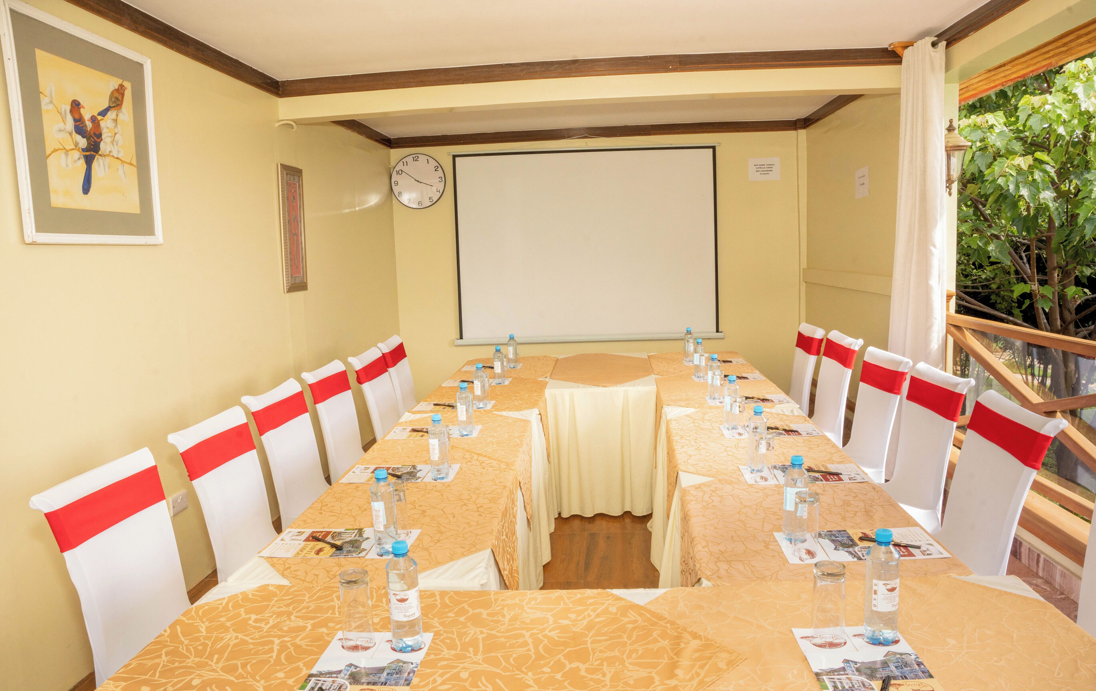 Un salón de conferencias con una larga mesa, sillas adornadas con cintas rojas, botellas de agua, un reloj y una pantalla para proyector.