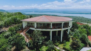 A large, circular building with a red roof, surrounded by lush greenery and a lake in the background.