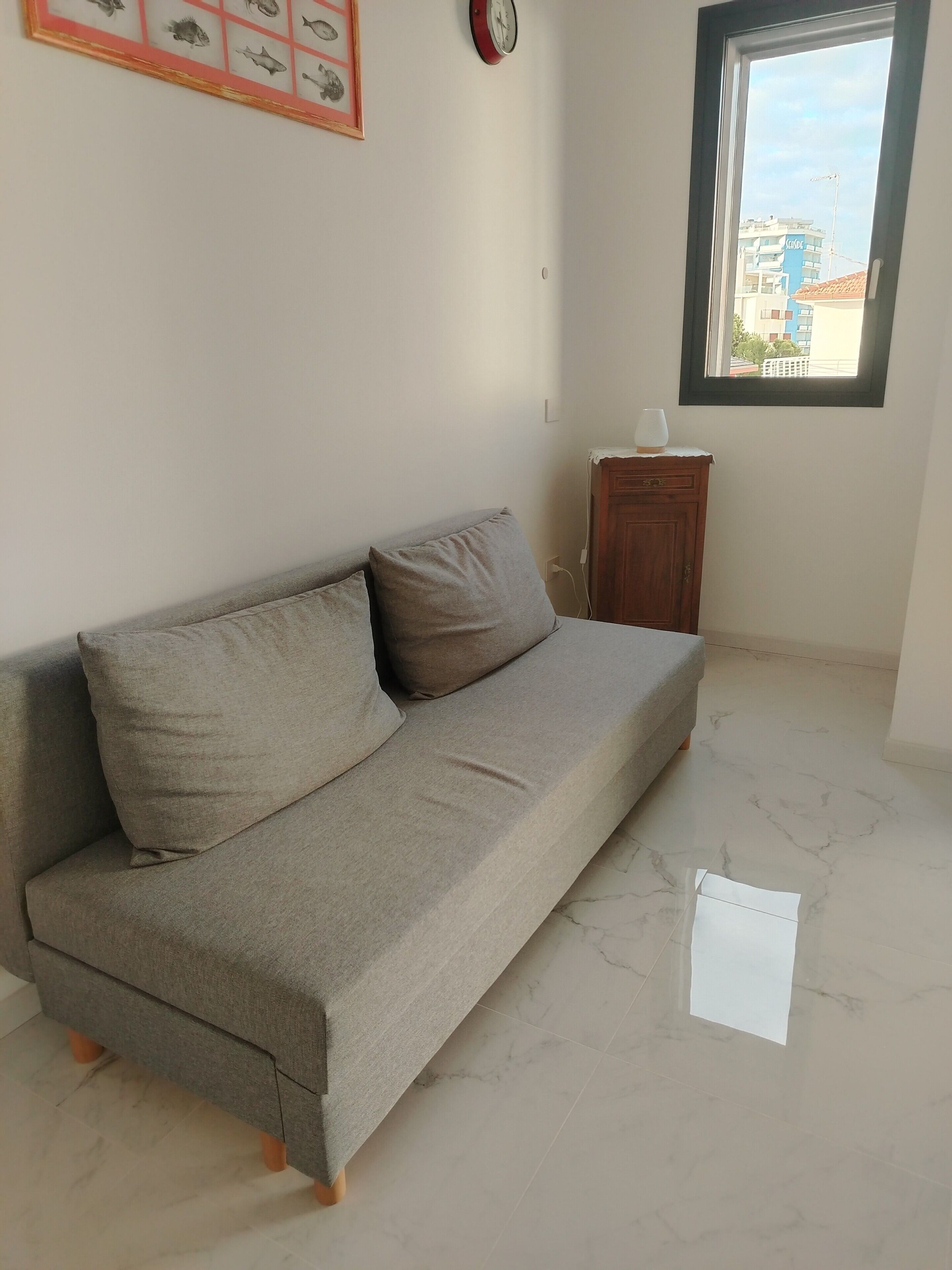 Una habitación con un sofá gris, un reloj, un cuadro de peces enmarco, una ventana y un armario de madera.