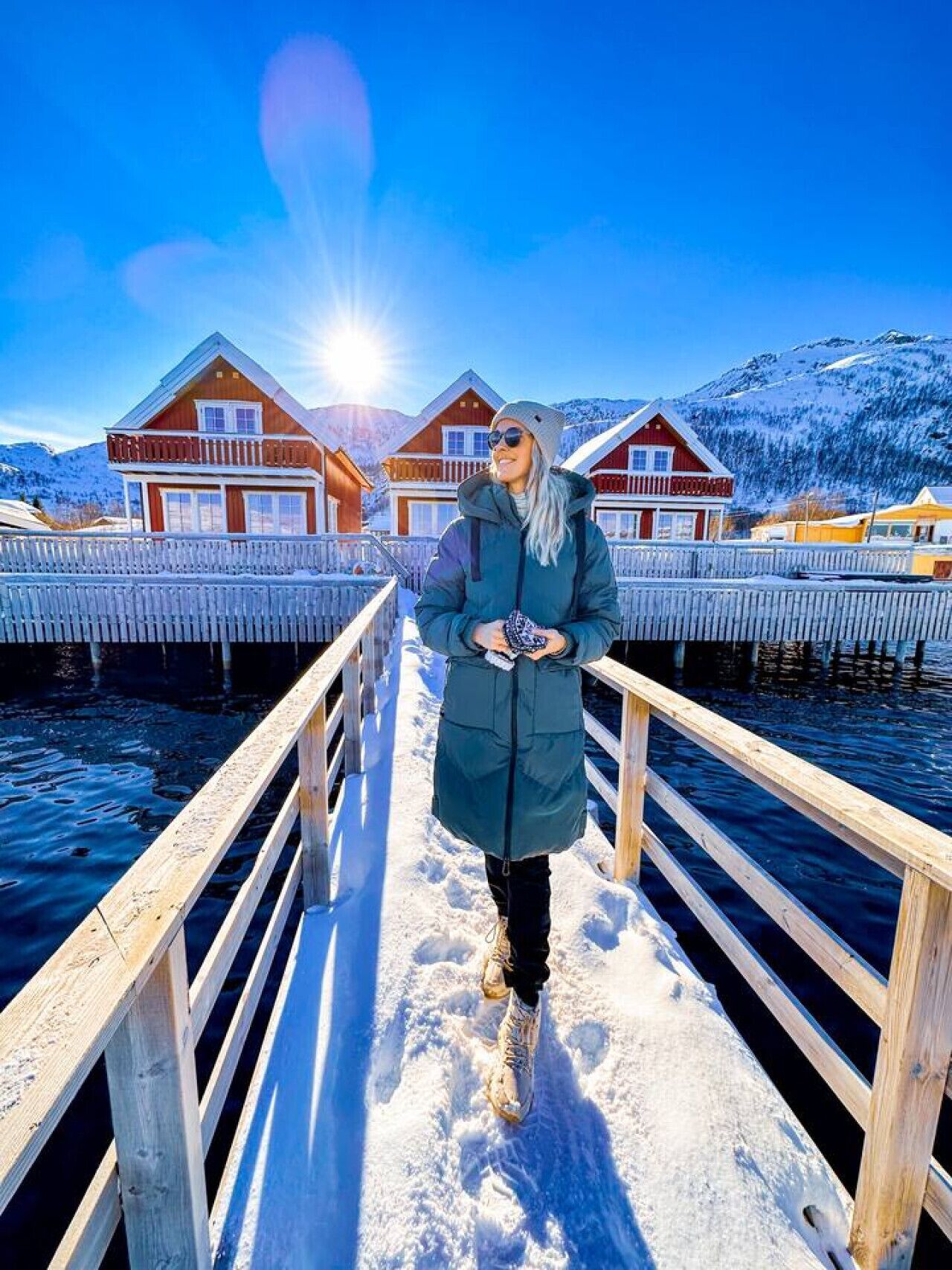 A person standing on a wooden pier with snow-covered ground, overlooking a fjord with wooden houses and mountains in the background.