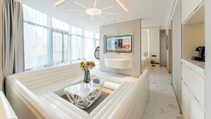 A modern hotel room with a white sofa, a coffee table, a TV, and a large window with curtains.
