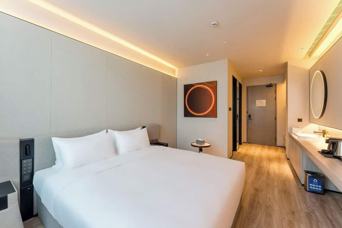 Una habitación de hotel moderna con una cama grande, panel de control junto a la cama, un espejo redondo y una pequeña mesa con una taza.