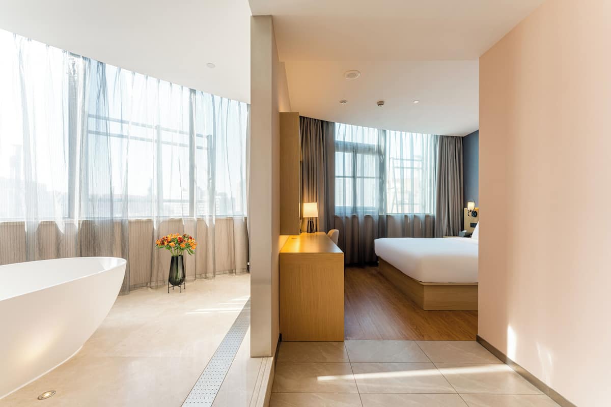 Una habitación de hotel moderna con una bañera grande, un escritorio pequeño, una cama y ventanas amplias con cortinas ligeras.