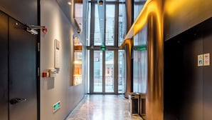 A modern hotel corridor with a glass elevator, a fire alarm, and a visible exit sign.