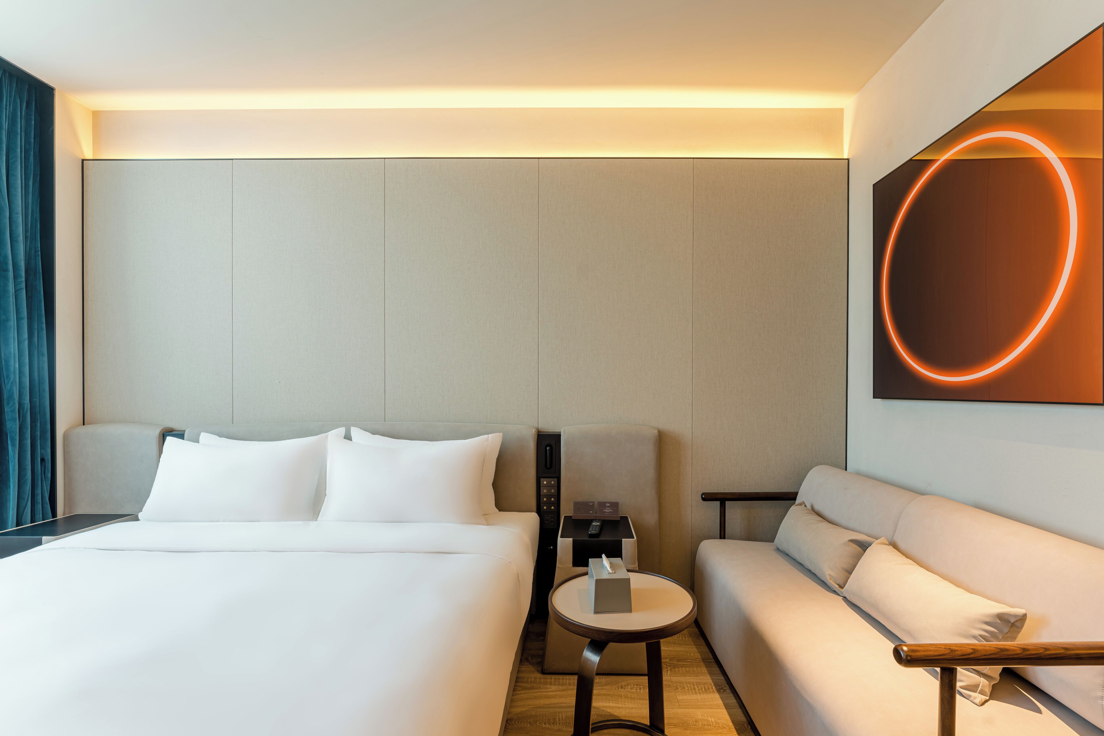 A modern hotel room with a bed, a sofa, a small table, and a wall-mounted light fixture.