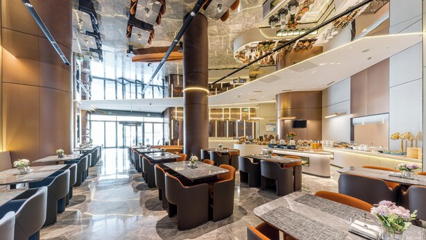 A modern hotel lobby with a central column, multiple seating areas, a reception desk, and a view of the upper floor.