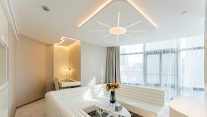 Een moderne hotelkamer met een witte bank, een bijzondere salontafel, een vaas met bloemen, een groot raam met gordijnen en een plafondlamp.