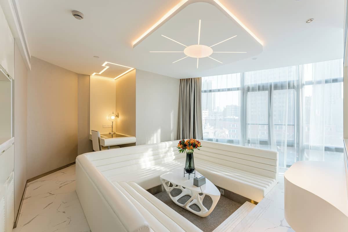 Una habitación de hotel moderna con un sofá blanco, una mesa de centro original, un jarrón con flores, un ventanal con cortinas y una lámpara en el techo.