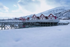 Række røde træhuse med en træbro, der strækker sig ud i en frossen sø, omgivet af sneklædte bjerge.
