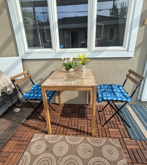 Uma mesa de madeira com duas cadeiras azuis com bolinhas, uma planta em vaso e flores amarelas num pátio com chão em padrão herringbone.