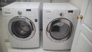 Two white front-loading washing machines with control panels, placed side by side in a room with a tiled floor. - Sunshine Home Vancouver Near UBC/DT/YVR  Family-sized Studio (Vancouver)