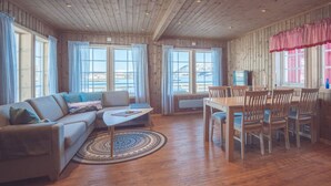 Un salon chaleureux avec un canapé, une table basse et une table à manger avec des chaises. De grandes fenêtres sont ornées de rideaux, le plafond est en bois et on peut profiter d’une vue sur l’extérieur.