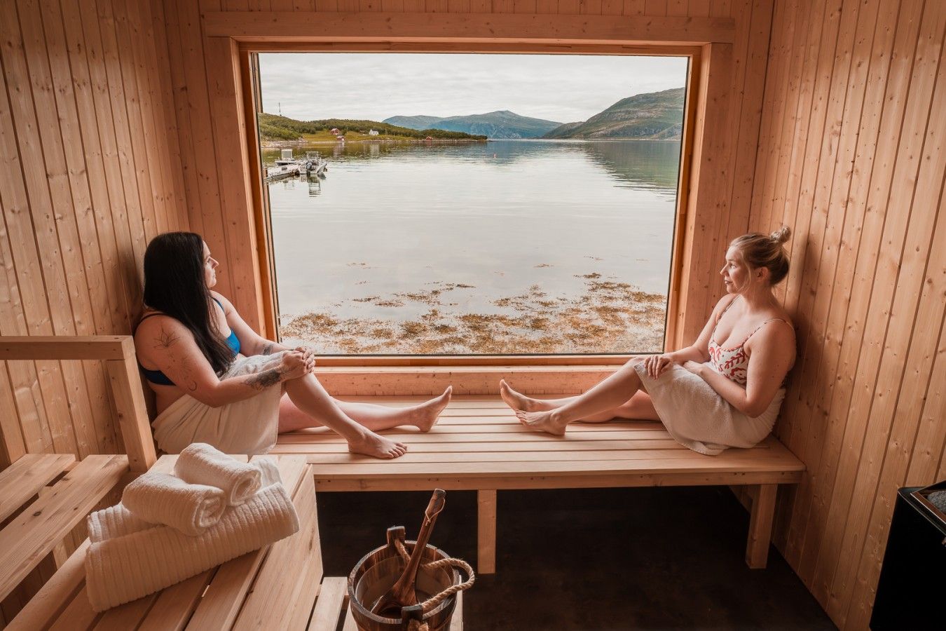 Two individuals seated on wooden benches in a sauna room with a large window overlooking a calm lake and hills.