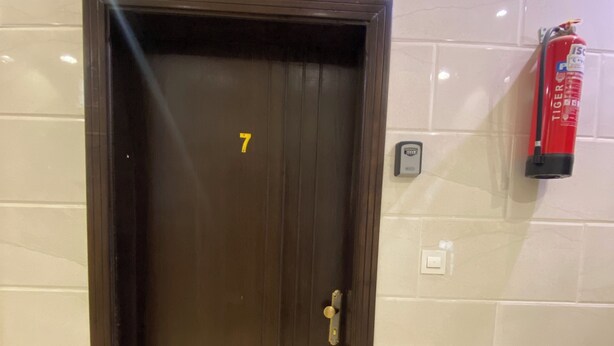 Una puerta marrón con el número 7, un extintor y una lámpara fijada a la pared.