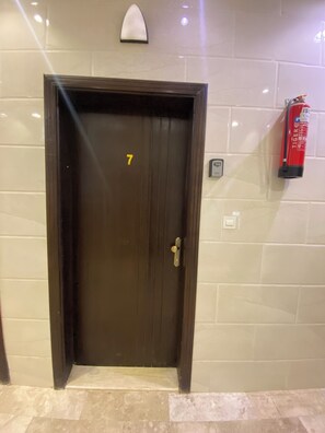 A brown door with the number 7, a fire extinguisher, and a wall-mounted light fixture. - Avenor Apartment J7 – Spacious 1-Bedroom Apartment with Private Bathroom | Al Hamdaniyah (Jeddah)