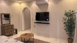 Una habitación de hotel moderna con una televisión de pantalla plana montada en la pared, un aparador de madera, una mesita con una bandeja, una planta en maceta y un espejo grande.