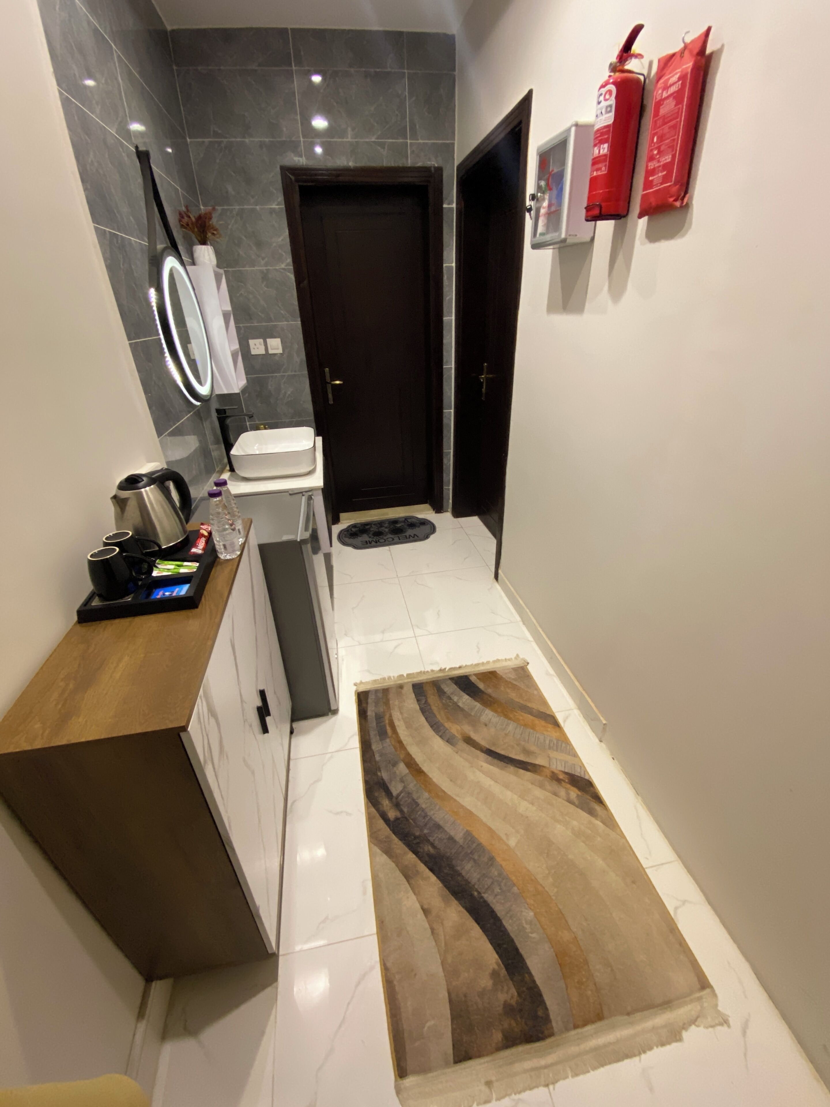 A modern kitchen with a coffee machine, a kettle, and a sink. There is a wooden counter and a white cabinet. A rug is on the floor, and a door is visible in the background.