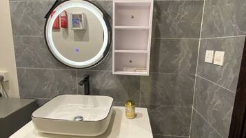 Un baño con tocador blanco, un espejo redondo, un estante blanco pequeño y un bote de basura rojo.