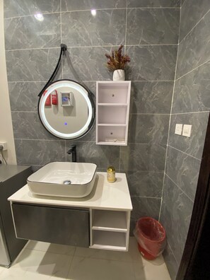 A bathroom with a white vanity, a round mirror, a small white shelf, and a red trash bin. - Avenor Apartment J7 – Spacious 1-Bedroom Apartment with Private Bathroom | Al Hamdaniyah (Jeddah)