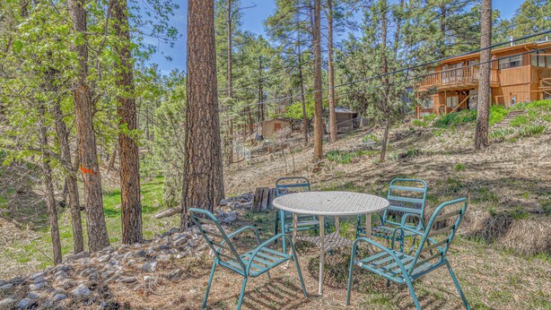 A rustic outdoor seating area with metal chairs and a table, surrounded by tall pine trees and a wooden building in the background. - Great for Couples! Studio Casita in Ruidoso (Ruidoso)