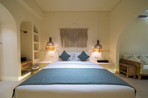 A hotel room with a large bed, two bedside lamps, a wall-mounted macrame piece, and a seating area with a sofa and chairs. - Velin Villa (Canggu)