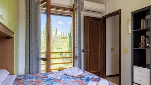 Un dormitorio con una cama, una estantería y una puerta que da a un balcón con vistas a árboles y colinas.