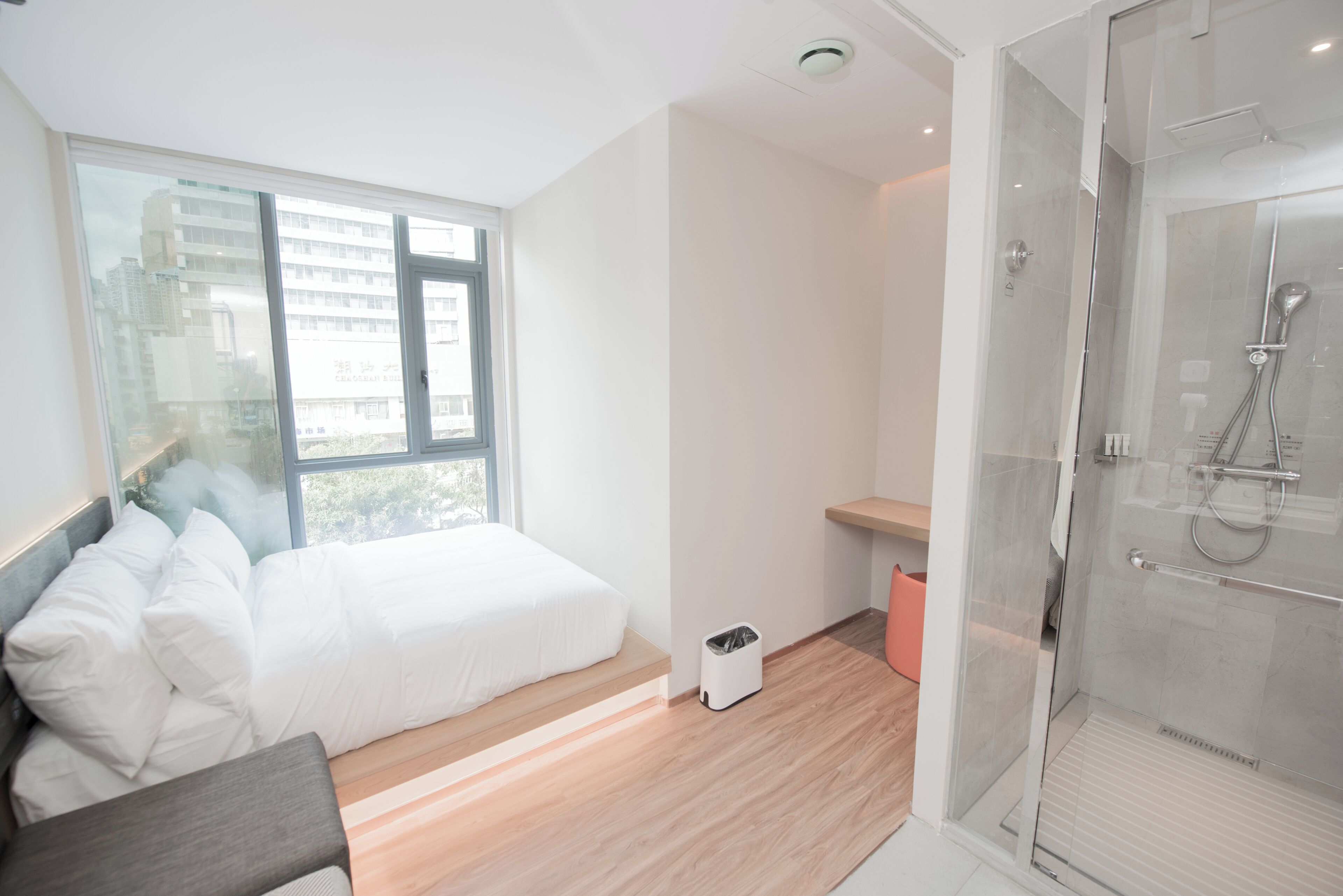 A modern hotel room with a large window, a bed, a shower area, and a small trash bin.