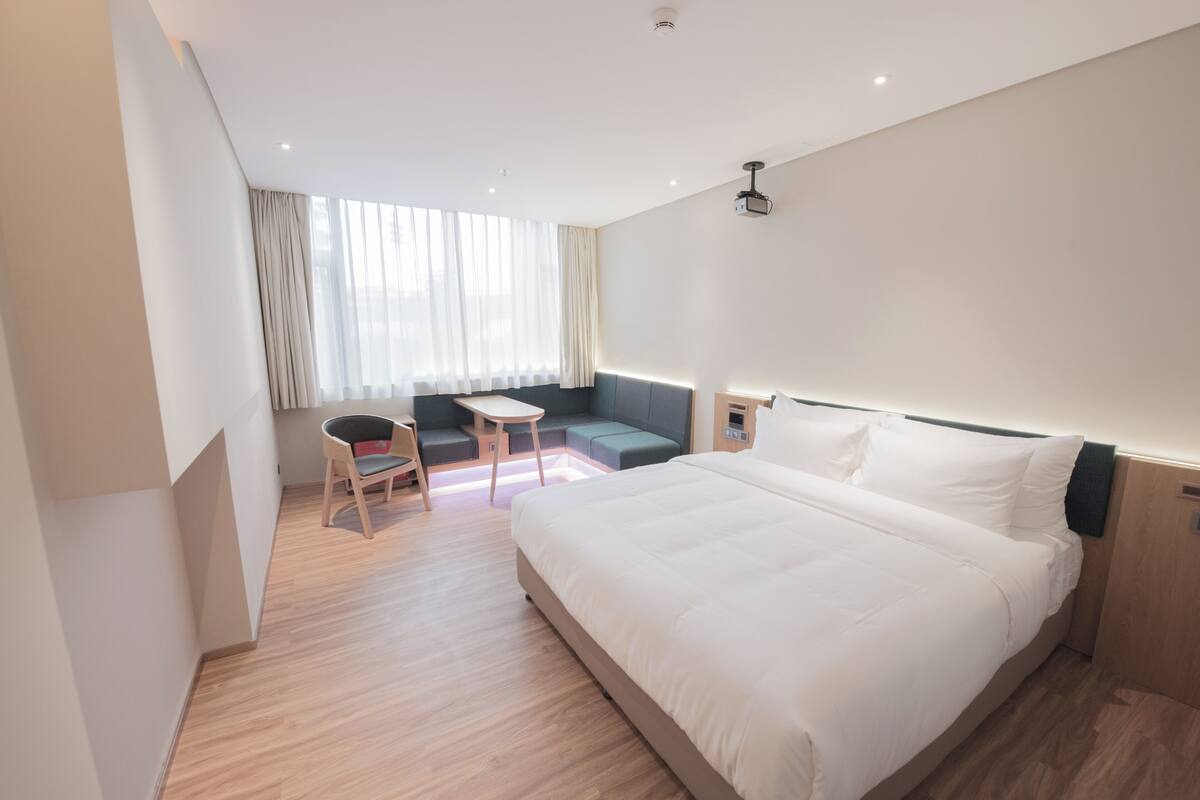 Una habitación de hotel moderna con una cama grande, un rincón de descanso pequeño y un ventanal con cortinas.