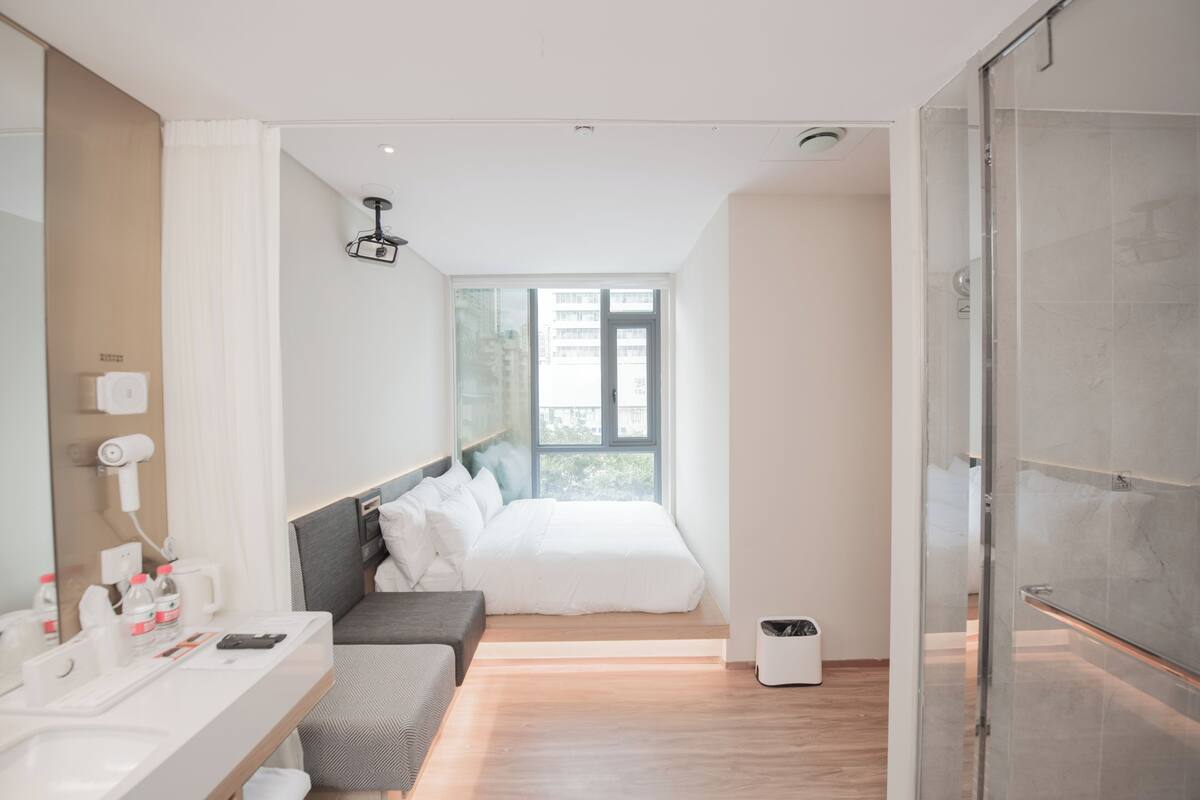 Una habitación de hotel moderna con cama, mesita de noche, espejo, ducha y ventana.
