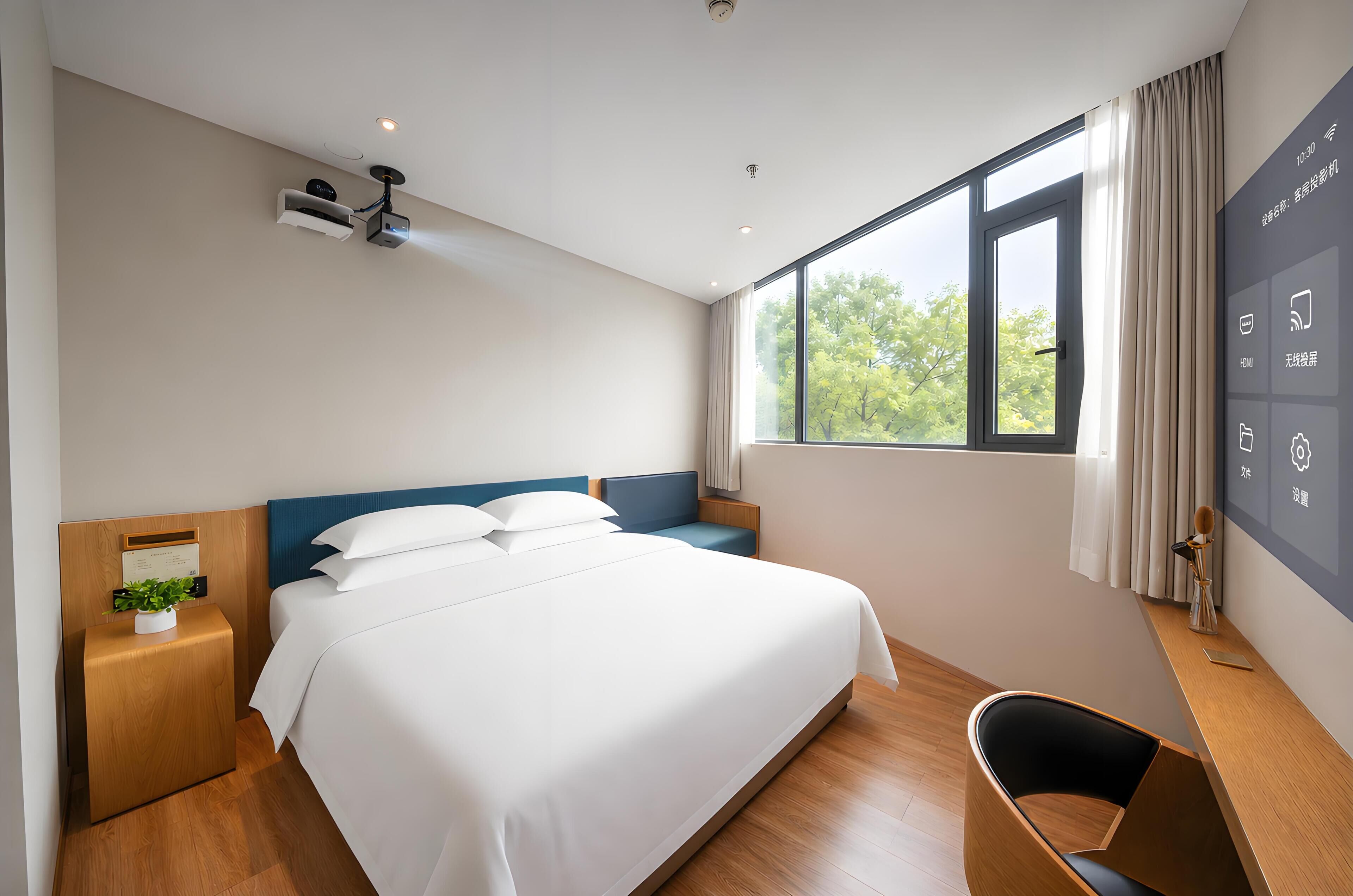 A modern hotel room with a large bed, a desk, a chair, and a window with a view of trees.