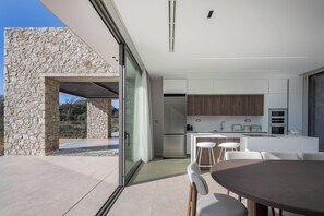 A modern kitchen with a stone exterior, a dining area with a round table, and a view of the outdoors. - Aeora Villa, Shoreline Getaway (Chania)