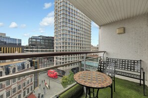 Ein Balkon mit einem karierten Tisch und einer schwarzen Metallbank, der auf eine Stadtstraße mit einem roten Bus und modernen Gebäuden blickt.