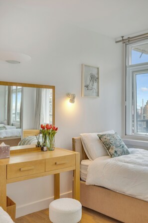 Ein Schlafzimmer mit Bett, Nachttisch, Spiegel, Fenster und Blick auf die Stadt.