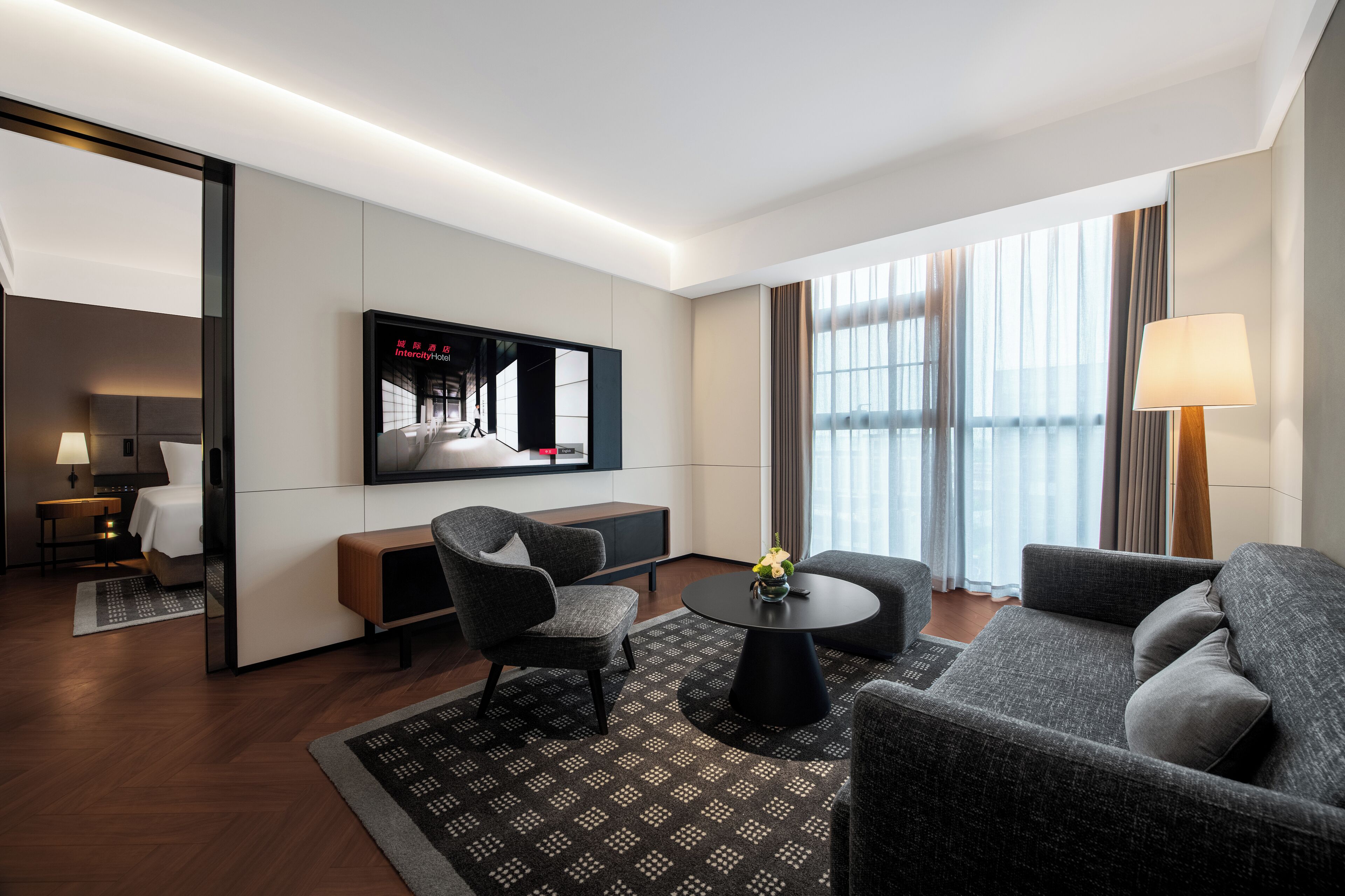 A modern hotel room with a sofa, armchair, round table, and a large flat-screen TV.
