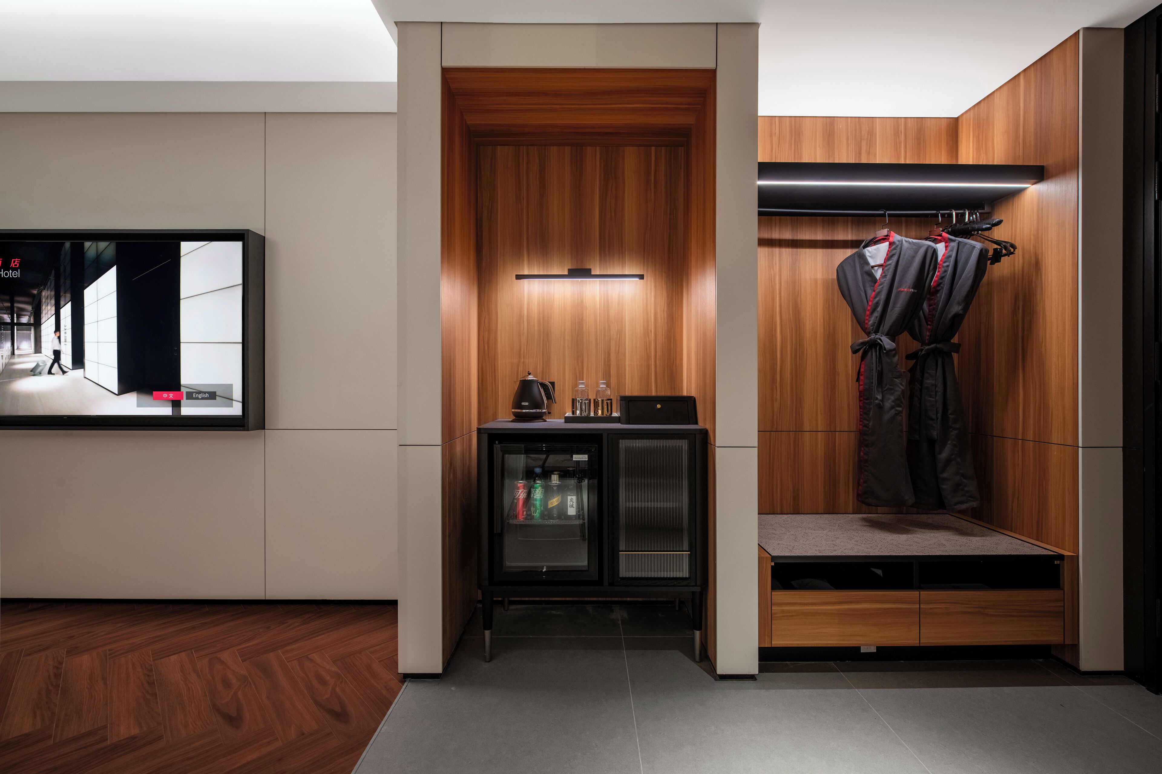 A modern hotel room with a wooden accent wall, a small seating area, a minibar, and a coat rack.