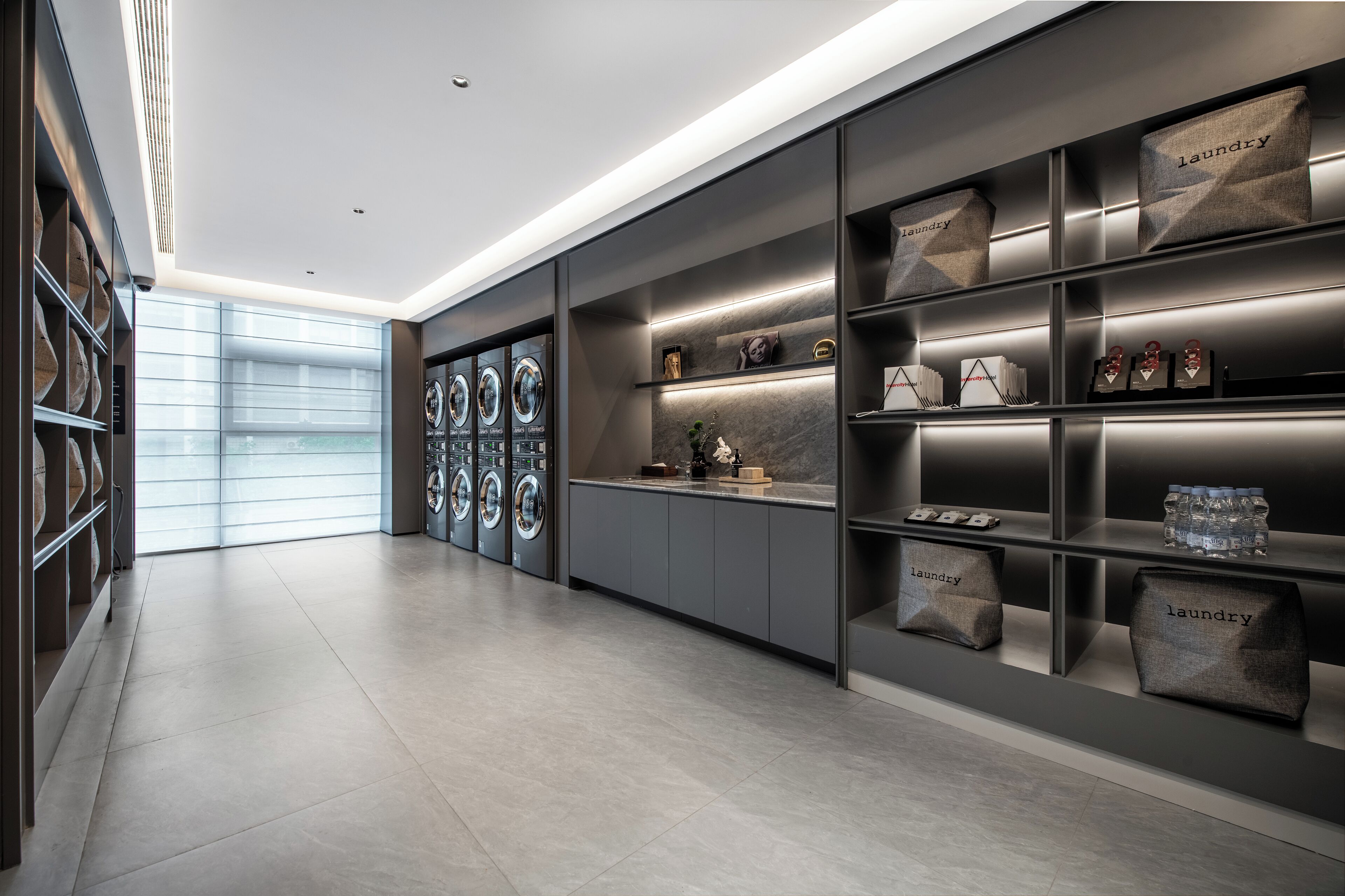 A modern laundry area with built-in washers and dryers, neatly organized shelves, and a large window providing natural light.