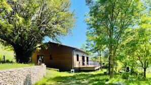 Un edificio rustico in legno con un muro in pietra e un ponte di legno, immerso nel verde e circondato da alberi.