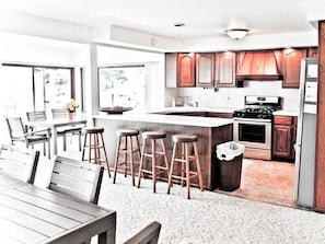 A modern kitchen with wooden cabinets, a central island with stools, a dining area with a table and chairs, and large windows.