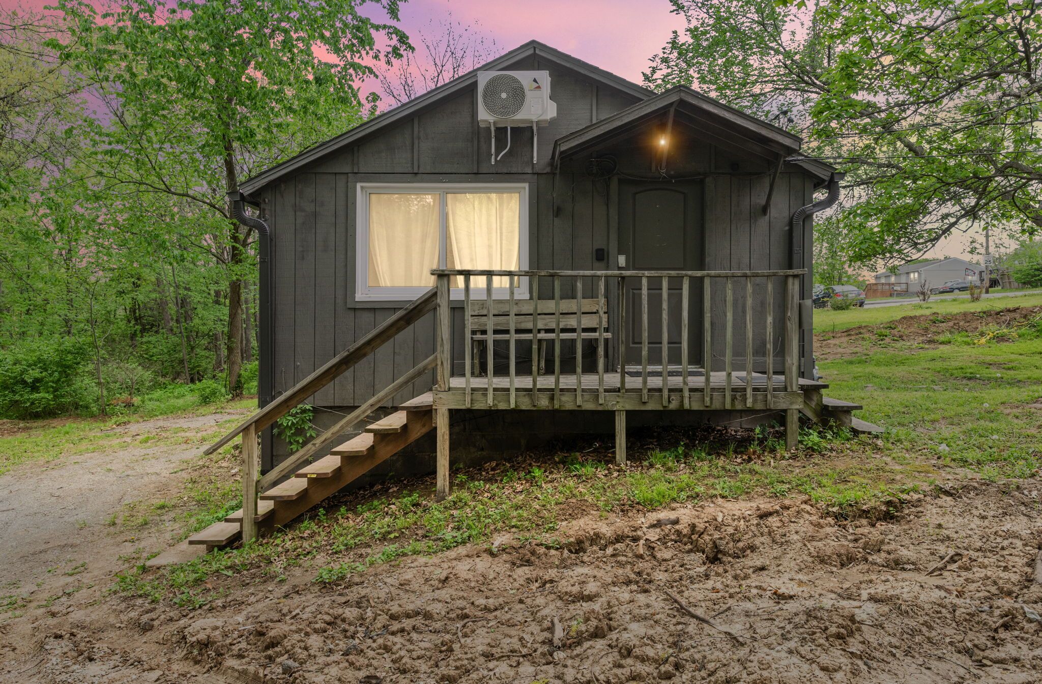 A small cabin with a wooden staircase leading to a porch, surrounded by trees and a dirt path.